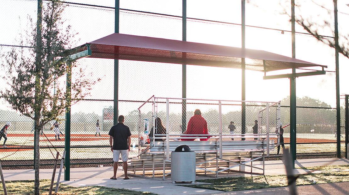 Red cantilever shade over aluminum bleachers beside a ball field