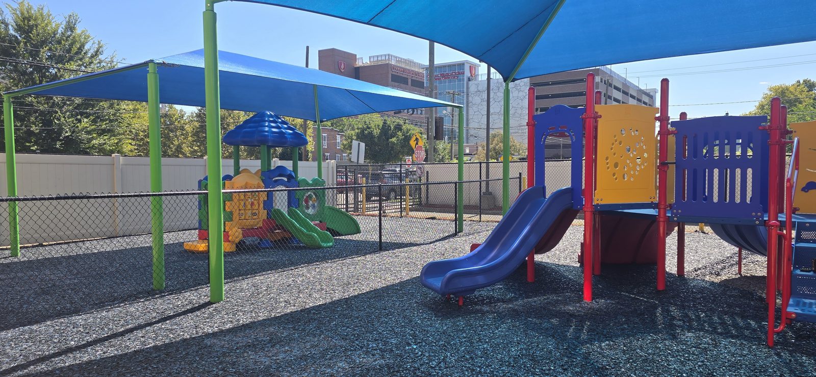 Two play structures under blue shade sails on dark bonded rubber surfacing at El Centro daycare in Camden, NJ, a fenced toddler unit on the left and a larger structure with a slide on the right