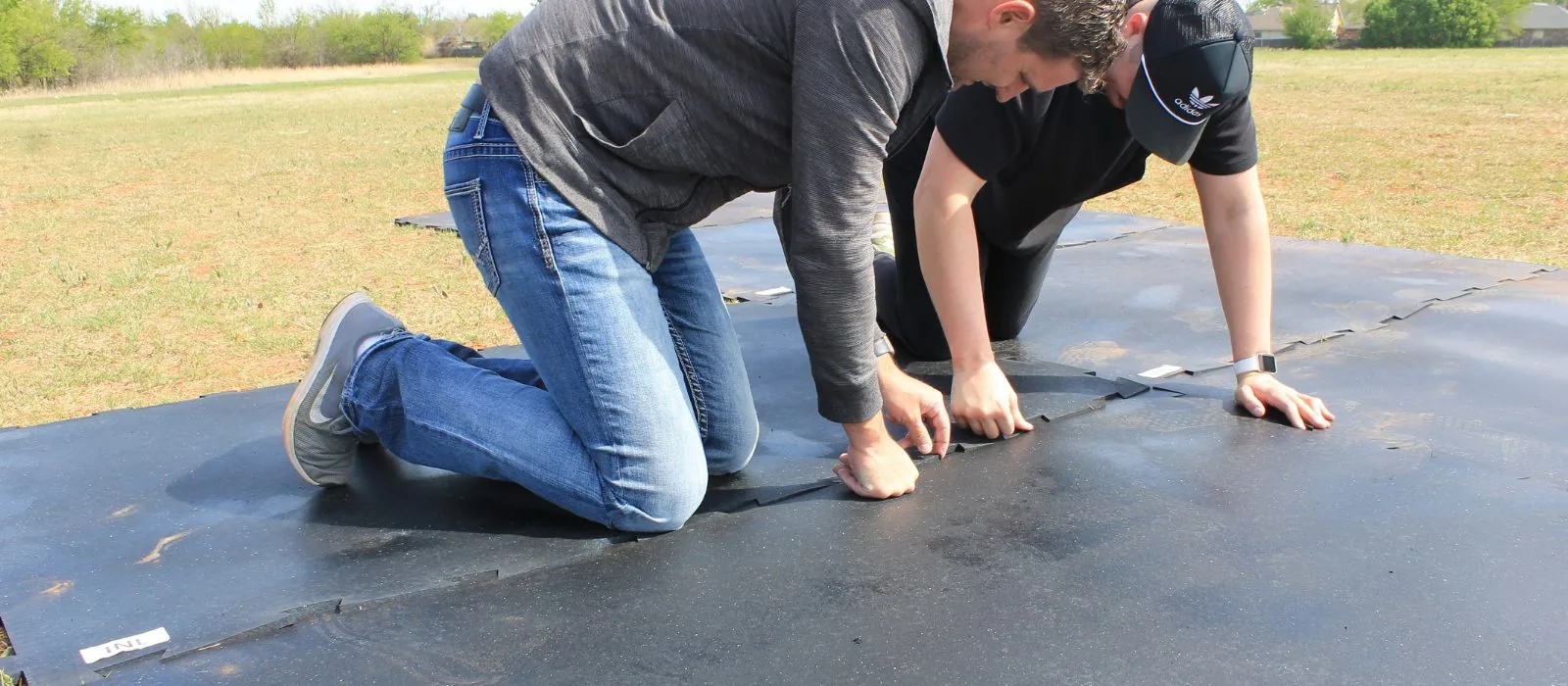 Two workers kneeling on grass, fitting interlocking black rubber floor mats together