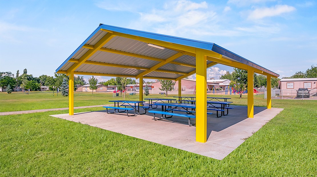 Gable-end steel shelter with a blue metal roof and yellow columns over blue picnic tables on a concrete pad