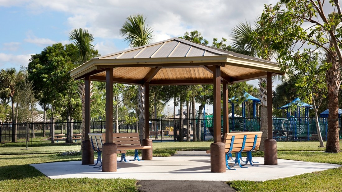 Hexagon steel shelter with a metal roof and brown columns over park benches on a concrete pad