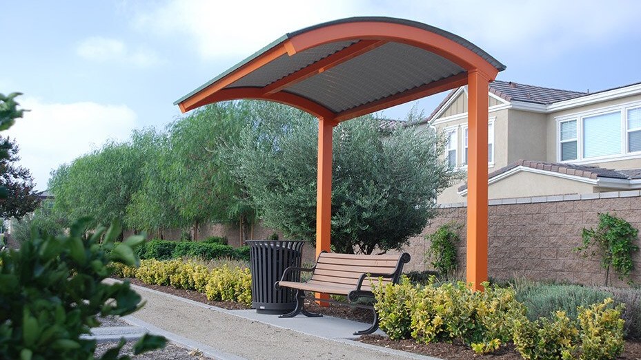 Arched cantilever steel shelter with orange columns on one side only, covering a bench beside a walkway