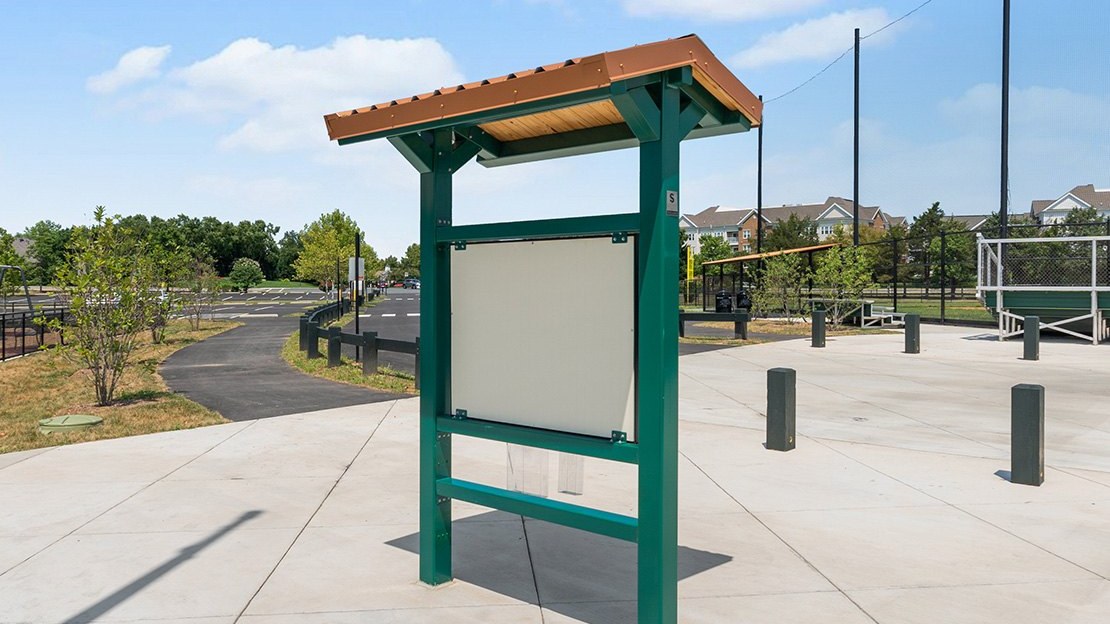 Green steel kiosk with a small metal roof over a blank display panel on a concrete plaza