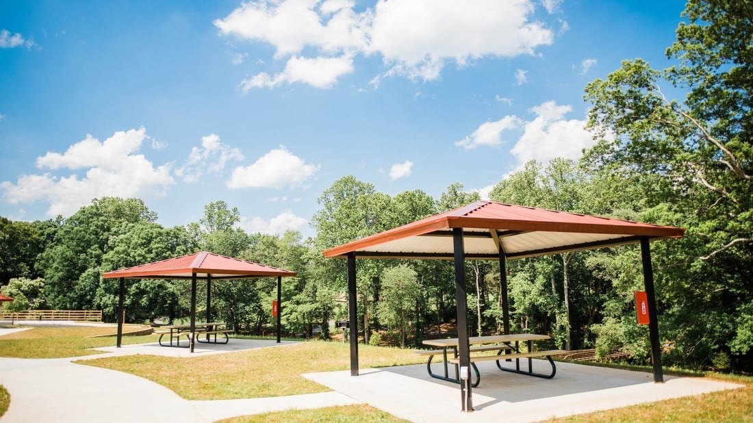 Two square steel shelters with red metal roofs and black columns over picnic tables on concrete pads beside a park path